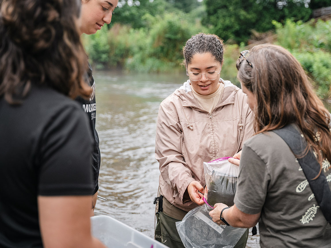 A college student in a tan jacket collects samples in a stream